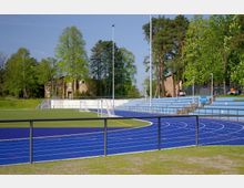 Sportanlage mit einer blauen Laufbahn, einem angrenzenden Kunstrasenfeld und mehreren weißen Toren. Im Hintergrund sind Bäume, Sitztribünen und ein Backsteingebäude sichtbar.