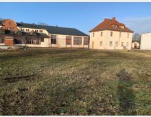 Ein offenes Gelände mit einer Wiese im Vordergrund, im Hintergrund ein helles zweistöckiges Gebäude mit rotem Ziegeldach und daneben ein größeres Industriedachgebäude mit großen Fensterfronten. Der Himmel ist blau, und das Gelände wirkt teils brachliegend.