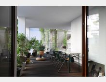 Überdachte Terrasse mit Holzfußboden, dekoriert mit mehreren Pflanzen in Töpfen. Auf der Terrasse stehen ein Esstisch mit Stühlen, zwei Liegestühle sowie ein kleiner Rattanhocker, mit sichtbarer Begrünung an den Geländern.