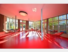 Moderner Innenraum mit rotem Boden, großen Fenstern und Gartenblick. Der Raum ist mit runden Metallstühlen, Vorhängen und einer zentralen Deckenlampe ausgestattet.