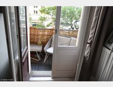 Blick durch eine geöffnete Balkontür auf einen kleinen Balkon mit Bambusmatten als Sichtschutz, einem hellgrauen Sofa mit Kissen, einem kleinen Holztisch und einem Glaswindlicht. Im Hintergrund sind Bäume und ein Gebäude erkennbar.