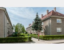 Mehrfamilienhäuser in einem Wohngebiet mit schlichten, hellbraunen Fassaden und roten Ziegeldächern. Im Vordergrund befinden sich gepflegte Hecken, ein Fußweg und ein einzelner Nadelbaum, umgeben von einer grünen Umgebung.