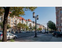 Straßenszene mit mehrstöckigen Wohn- und Geschäftshäusern in verschiedenen Farben, angrenzend an eine von Bäumen gesäumte Hauptstraße mit parkenden Autos und Fußgängerwegen. Im Vordergrund stehen Laternen und ein Verkehrsschild, rechts ist der Eckbereich eines Gebäudes sichtbar.