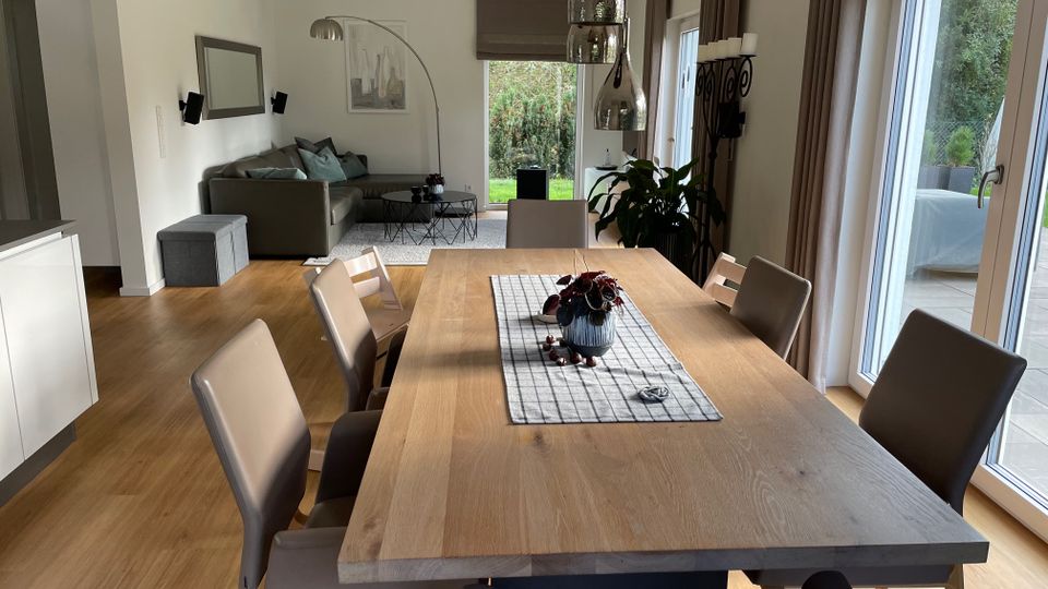 Essbereich und Wohnzimmer in einem modernen Haus mit hellem Holzboden, einem Esstisch aus Holz und gepolsterten Stühlen. Große Fenster lassen viel Tageslicht herein, im Hintergrund befindet sich eine graue Couch und eine Stehleuchte.