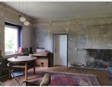 Ein schlicht eingerichteter Raum mit abgeblätterten Wänden, einem Holzfußboden und einem bunten Orientteppich. Im Raum stehen ein runder Holztisch mit einem Klappstuhl, ein elektronisches Keyboard, und ein Fenster lässt Tageslicht herein.