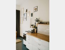 Helle Küche mit weißen Schränken und einer Arbeitsplatte aus Holz. Im Hintergrund befinden sich ein weißer Herd, dekorative Wandbilder, Pflanzen und Gläser auf einem kleinen Regal vor einer weißen Fliesenwand.