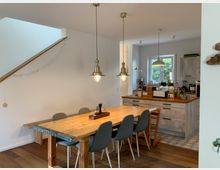 Wohnküche mit Holzboden, einem großen Esstisch aus Holz und sechs Stühlen. Im Hintergrund befindet sich eine Küche mit weißen Schränken, hölzernen Arbeitsplatten, einer Fensterfront und mehreren Hängelampen.