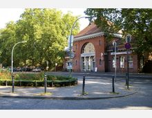 Backsteingebäude eines Bahnhofs mit rundem Fenster über dem Eingang, auf dem "Sonnenallee" steht. Im Vordergrund befinden sich ein Kreisverkehr mit Blumenbeet, Straßenlaternen und Fußwege, im Hintergrund große Bäume.
