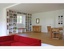 Wohnzimmer mit hellem Holzfußboden, einer roten Sofaecke im Vordergrund und einer weißen Wand mit integrierten Bücherregalen sowie einem Fenster. Im Hintergrund befinden sich ein Holztisch mit Stühlen, eine kleine Kommode und eine Tür.