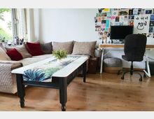 Wohnzimmer mit hellbraunem Sofa, roten und beigen Kissen sowie einem niedrigen Couchtisch mit Tischläufer und Pflanzenarrangement. Im Hintergrund steht ein Schreibtisch mit Computer, schwarzem Büro-Stuhl und an der Wand eine Collage aus Postkarten und Fotos.