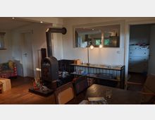 Gemütlicher Wohnbereich mit einem Holzofen in der Mitte, flankiert von einem Regal mit Büchern und Kisten. Der Raum verfügt über Holzböden, einen Esstisch und eine offene Durchreiche, die Einblick in eine Küche mit modernen Geräten gibt.