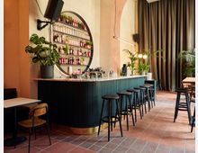 Ein stilvoller Barbereich mit grünem Tresen, schwarzen Hockern und einer wandmontierten, runden Regalstruktur mit Flaschen. Der Raum ist dekoriert mit großen Zimmerpflanzen, hohen Vorhängen und warmen Wandfarben.