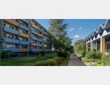 Mehrstöckiges Wohngebäude mit bunten Balkonen auf der linken Seite, daneben ein schmaler gepflasterter Weg und mehrere Grünflächen mit Bäumen und Sträuchern; rechts ein modernes Gebäude mit sägezahnförmigem Dach.