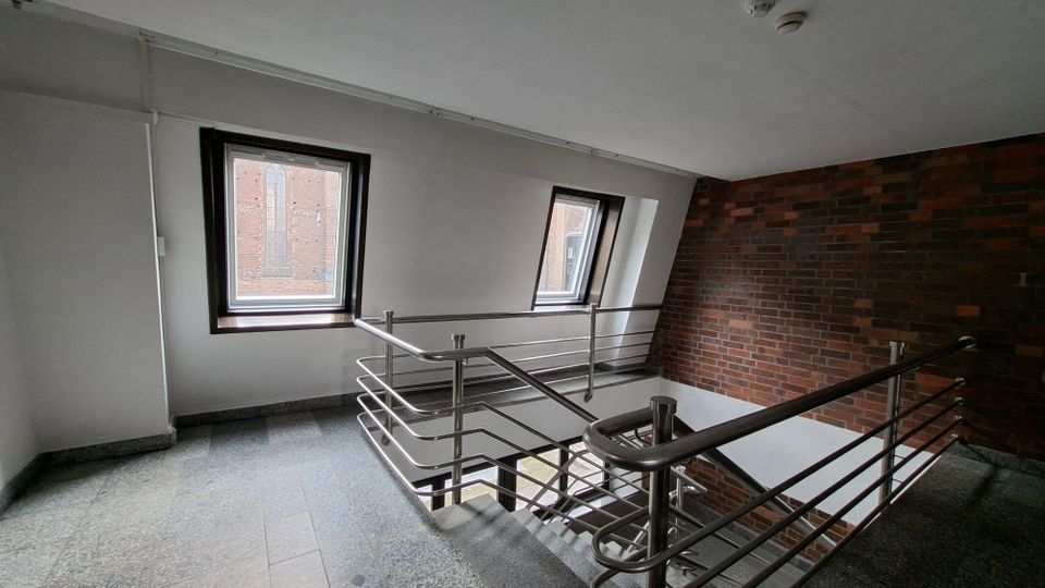 Innenliegendes Treppenhaus mit grauem Steinboden und Metallgeländer, daneben zwei rechteckige Fenster in einer schrägen Wand mit Blick auf eine Backsteinfassade. Eine Backsteinwand verläuft entlang des Treppenabgangs unter einer weißen Decke.