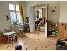 Ein Kinderzimmer mit Holzboden, einem kleinen Tisch mit Stühlen und einem Schaukelpferd im Vordergrund. Im Hintergrund führt eine doppelte Holztür zu einem weiteren Raum mit Spielzeug, einem großen Plüschpferd und Regalen voller Bücher und Spielsachen.