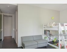 Kinderzimmer mit schlichtem Design, bestehend aus einem weißen Hochbett mit Regal darunter, einer grauen Couch und Spielzeug sowie Büchern auf dem Boden. Helle Wände und dunkler Holzboden sorgen für einen minimalistischen Eindruck.