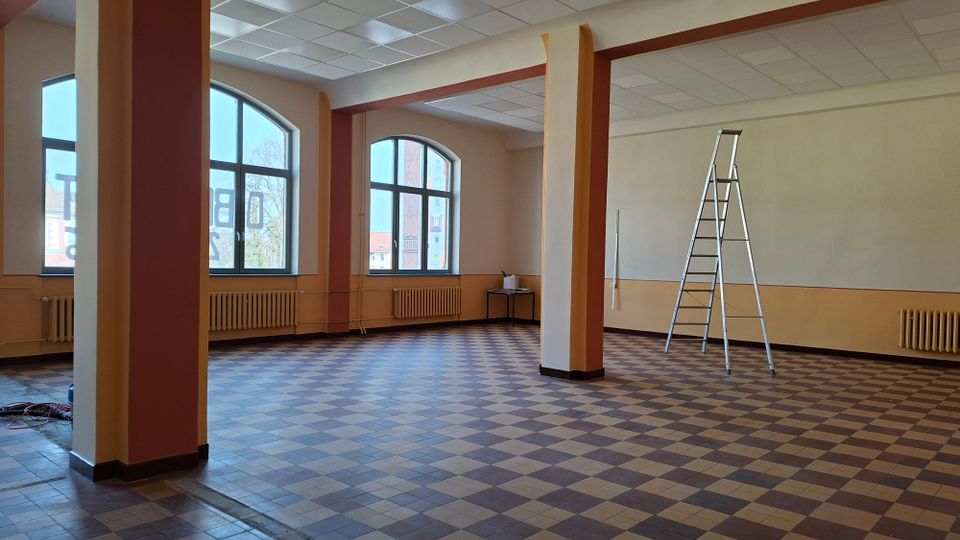 Großer, leerer Innenraum mit kariertem Fliesenboden, mehreren tragenden Säulen und einer abgehängten Rasterdecke. An der Außenwand befinden sich große Rundbogenfenster mit Heizkörpern darunter; rechts steht eine aufgeklappte Stehleiter.