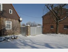 Verschneiter Hof zwischen einem Holzhaus mit Satteldach und einem Backsteingebäude, getrennt durch ein hohes Metalltor und einen gemauerten Torpfeiler. Im Vordergrund liegt Schnee, rechts steht ein kahler Baum vor der Backsteinfassade unter klarem Himmel.