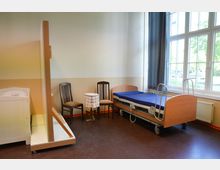 Zimmer mit einem Pflegebett auf Rollen, zwei Holzstühlen und einem kleinen Tisch mit Spitzendecke vor einem Fenster mit Vorhängen. Eine Trennwand teilt den Raum, auf der linken Seite ist ein weißes Babybett erkennbar.