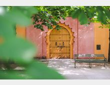 Rosa-orangefarbene Wand mit einem gelben Holztor in einem arabischen Bogenstil, umgeben von Bäumen, deren Blätter teilweise den Blick verdecken; im Vordergrund steht rechts eine grüne Parkbank.