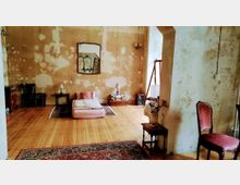 Ein minimalistischer Innenraum mit abgenutzten Wänden, Holzboden und spärlicher Einrichtung, darunter eine niedrige Matratze, ein Spiegel an der Wand, mehrere kleine Lampen sowie vereinzelte Möbelstücke wie ein Stuhl und ein kleiner Beistelltisch.