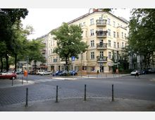 Straßenszene mit einem hellgelben Eckgebäude im Jugendstil mit verzierten Balkonen und Geschäften im Erdgeschoss. Im Vordergrund sind eine Kopfsteinpflasterstraße, parkende Autos und vereinzelt Bäume zu sehen.