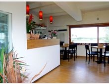 Modernes Café mit hellen Wänden, Holzboden und großen Fenstern mit Blick auf eine grüne Landschaft. Es gibt einen Tresen mit Dekorationen und mehrere einfache schwarze Tische und Stühle.