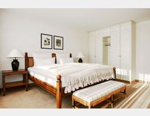 Helles Schlafzimmer mit einem großen Holzbett, flankiert von zwei Nachttischen mit Lampen. Im Hintergrund ein weißer Einbauschrank mit goldenen Griffen und zwei Wandbilder über dem Bett, im Vordergrund eine gepolsterte Sitzbank.