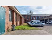 Industriehof mit roten Backsteingebäuden, großen Fenstern und einem begrünten Rasenstreifen. Im Vordergrund parken mehrere Fahrzeuge, darunter Lieferwagen und ein Pkw, daneben ein offener Unterstand mit weiteren geparkten Fahrzeugen.