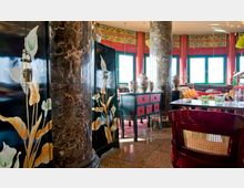 Innenraum eines asiatisch inspirierten Restaurants mit dekorativen schwarzen Möbelstücken, die mit floralen Mustern verziert sind. Dunkle Marmorsäulen, leuchtend rote und grüne Akzente, traditionelle Keramikvasen und große Fenster mit Blick auf Tageslicht bestimmen das Ambiente.