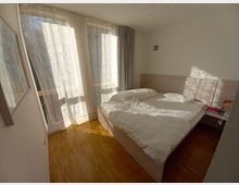 Ein helles Schlafzimmer mit einem Doppelbett, weißen Bettbezügen und einem hellen Holzfußboden. Neben dem Bett befinden sich kleine Nachttische mit Büchern und einem Wecker, und durch bodentiefe Fenster mit gemusterten Vorhängen fällt Sonnenlicht in den Raum.