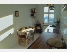 Helle Küche mit einem langen, hölzernen Arbeitsbereich auf der rechten Seite, einer kleinen Sitzgruppe mit Holztisch und Stühlen links und großen Fenstern im Hintergrund, die Tageslicht hereinlassen. An der Wand hängt ein Regal mit Utensilien, und darunter befinden sich weitere Küchenutensilien auf einem kleinen Holzcaddy.