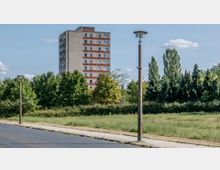 Wohnblock eines Plattenbaugebäudes mit hellgrauer und roter Fassade im Hintergrund, umgeben von Bäumen, Sträuchern und einer Wiese. Im Vordergrund sind eine asphaltierte Straße, ein Gehweg und zwei Straßenlaternen zu sehen.
