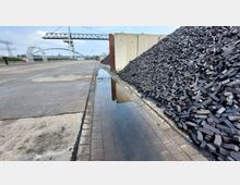 Industriegelände mit einer großen Stapelung von schwarzen Kohlebriketts rechts, einer Betonfläche links und einer Pfütze auf einem gepflasterten Weg in der Mitte. Im Hintergrund sind industrielle Strukturen wie ein Förderband und ein Bogenträger sichtbar.