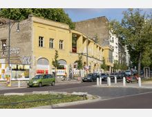 Straßenszene mit einem gelben, teilweise abgenutzten Gebäude mit Bögen und bröckelndem Putz; Plakate bedecken die Wände. Im Vordergrund sind parkende Autos, ein Motorrad und eine baumbestandene Straße zu sehen.