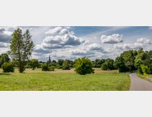Weite Grünfläche mit einzelnen Bäumen und Büschen, im Hintergrund eine Kirchturmspitze, umgeben von dichter Vegetation. Ein schmaler asphaltierter Weg führt durch die Landschaft, darüber ein Himmel mit vielen Wolken.