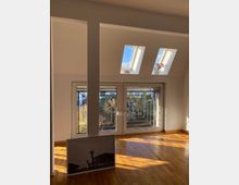 Wohnraum mit Holzboden, großen Fenstern mit metallenen Gittern und zwei Dachfenstern, durch die Tageslicht einfällt. Im Vordergrund lehnt ein gerahmtes Bild an der Wand, im Fensterbereich steht eine Vase mit Blumen.