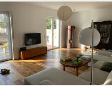 Helles Wohnzimmer mit Holzboden, großer Eckcouch und einem runden Couchtisch, auf dem Spielzeug liegt. Im Raum befinden sich ein Wandfernseher, ein Kaminofen in der Ecke, eine Bücherwand und bodentiefe Fenster mit Blick nach draußen.