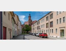 Eine gepflasterte Straße mit beidseitig stehenden Wohnhäusern führt zu einer Kirche im Hintergrund mit einem hohen Glockenturm aus Backstein. Vor den Häusern sind geparkte Autos und Laternen, daneben stehen grüne Bäume auf einem kleinen Platz.