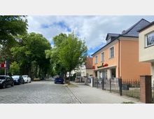 Wohnstraße mit Kopfsteinpflaster und parkenden Autos. Auf der rechten Seite steht ein zweistöckiges Gebäude mit orangefarbener Fassade, in dem sich eine Drogerie oder Apotheke befindet, während die Straße von Bäumen gesäumt ist.