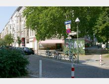 Straßenszene in einem urbanen Wohnviertel mit mehrstöckigen Altbauten auf der linken Seite, einem Gehweg mit Fahrrädern an Geländern befestigt und einer grünen Schautafel mit Aushängen. Im Hintergrund sind ein Laden mit rot-weiß gestreifter Markise und Bäume sichtbar.