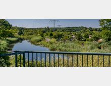Blick auf einen kleinen Fluss mit dichtem Schilf und umgebender Vegetation, im Hintergrund Gärten mit vereinzelten Hütten. Oberhalb verlaufen Stromleitungen, und am Horizont erstreckt sich ein Waldgebiet.