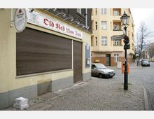 Straßenecke mit einem geschlossenen Laden mit der Aufschrift „Old Red Lion Inn“, daneben ein Laternenmast mit Straßenschild „Uthmannstraße“. Im Hintergrund stehen Wohngebäude mit gelber Fassade und geparkte Autos auf dem Kopfsteinpflaster.