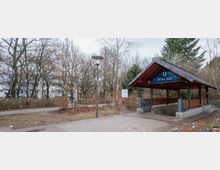 Außenansicht eines U-Bahnhofs mit der Beschriftung „Britz-Süd“ auf einem blauen Schild unter einem Holzdach. Der Eingang ist von Gehwegen, Bäumen und Sträuchern in einer urbanen Umgebung umgeben.