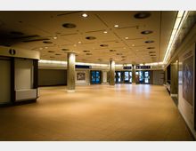 Heller, weitläufiger Innenbereich eines öffentlichen Gebäudes mit beigefarbenen Fliesenböden, mehreren beleuchteten Säulen und einer abgehängten Decke mit Lüftungsöffnungen. Im Hintergrund befinden sich Glastüren, Beschilderungen mit Richtungshinweisen sowie ein Informationsbereich.