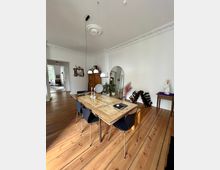 Esszimmer mit Holzboden und einem großen Esstisch aus recyceltem Holz, umgeben von schwarzen Stühlen. Im Raum befinden sich eine weiße Stuckdecke, eine Kommode, ein Spiegel, Pflanzen und ein Weinregal an der Wand.