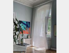 Helles Zimmer mit hohen Decken und verzierten Stuckdetails, einer geöffneten Balkontür mit leicht transparentem Vorhang, einem hölzernen Fischgrätboden, einem dekorativen Heizkörperdeckel und moderner Kunst an der Wand. Ein großer grüner Pflanzenzweig und ein aufgeschlagenes Buch liegen auf einem Tisch im Vordergrund.