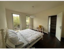 Schlafzimmer mit weißem Metallbett, dunklem Holzboden und zwei bodentiefen Fenstern, die auf einen Garten blicken. Im Hintergrund befindet sich ein kleiner Tisch mit gelbem Blumentopf sowie eine offene Tür zu einem angrenzenden Raum.