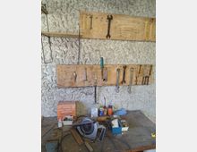 Werkstattwand aus rau verputztem Stein mit zwei Holzleisten als Werkzeughalter, an denen mehrere Schraubenschlüssel und Handwerkzeuge hängen. Davor eine staubige Werkbank mit verschiedenen Behältern, einer Kabelrolle und einer großen Metallsäge bzw. Schneidmaschine.