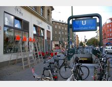 Straßenszene am U-Bahnhof Rosenthaler Platz in Berlin mit dem blauen U-Bahn-Schild, abgestellten Fahrrädern, einem weißen Gebäude mit großen Fenstern und mehreren geschäftigen Geschäften im Hintergrund. Eine Treppe führt in die U-Bahn-Station hinunter, während im Hintergrund Wohnhäuser und Straßenverkehr zu sehen sind.
